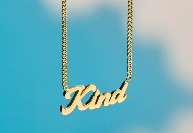 A blue sky background with clouds and a gold-tone La La Land x Fossil necklace that reads Kind.