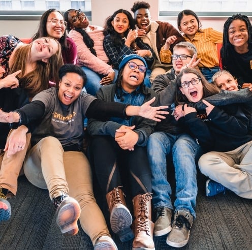 A large group of children sitting on the floor of a classroom smiling and hugging one another.