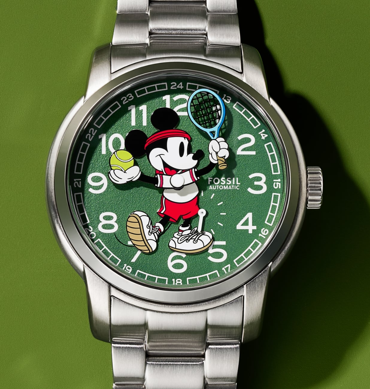Nuestro reloj Disney Mickey Mouse Tennis en edición limitada. Sobre una esfera con textura en color verde inspirada en la superficie de una pista de tenis, Mickey lleva un polo y unas zapatillas blancas, una bandana roja y unos pantalones cortos rojos. Sostiene una raqueta azul en una mano y una pelota de tenis en la otra mientras los brazos se mueven para contar las horas y los minutos.