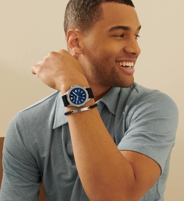 A man smiling as he looks over his shoulder. He’s wearing a light blue polo shirt, sporty watch with a dark blue dial and black leather bracelet with a silver detail.