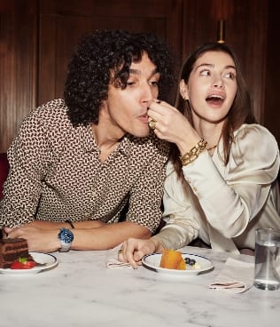 Une femme portant des bijoux dorés, faisant manger un gâteau à un homme portant la Fossil Blue GMT.