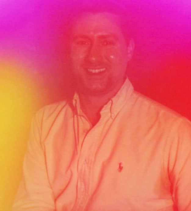 Fossil employee Drew, a Social Media Coordinator, is pictured smiling. The photo is overlaid with a rainbow filter effect to represent letting one’s Pride shine inside and out. It also includes the Fossil PLUS logo for LGBTQIA+ employees and allies, featuring two interlocking rainbows in the colours of the Pride and Trans flags.