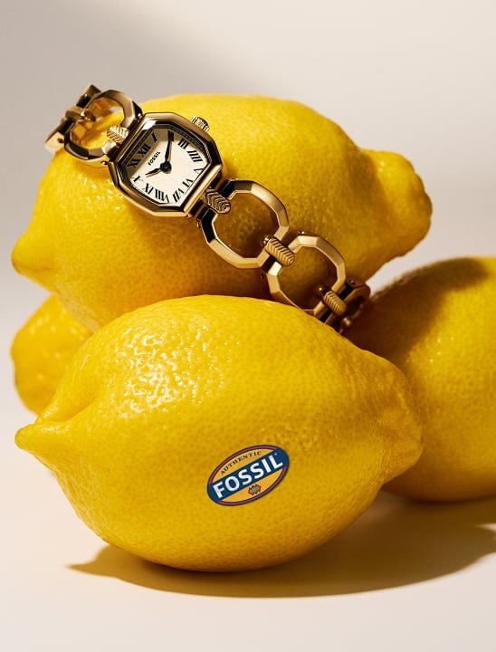 The Fossil Harlow mini chain watch is draped over a pile of ripe lemons with a Fossil sticker.