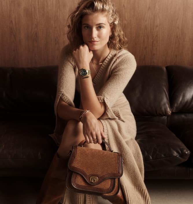 A woman sitting and holding a brown suede Fossil Heritage handbag.