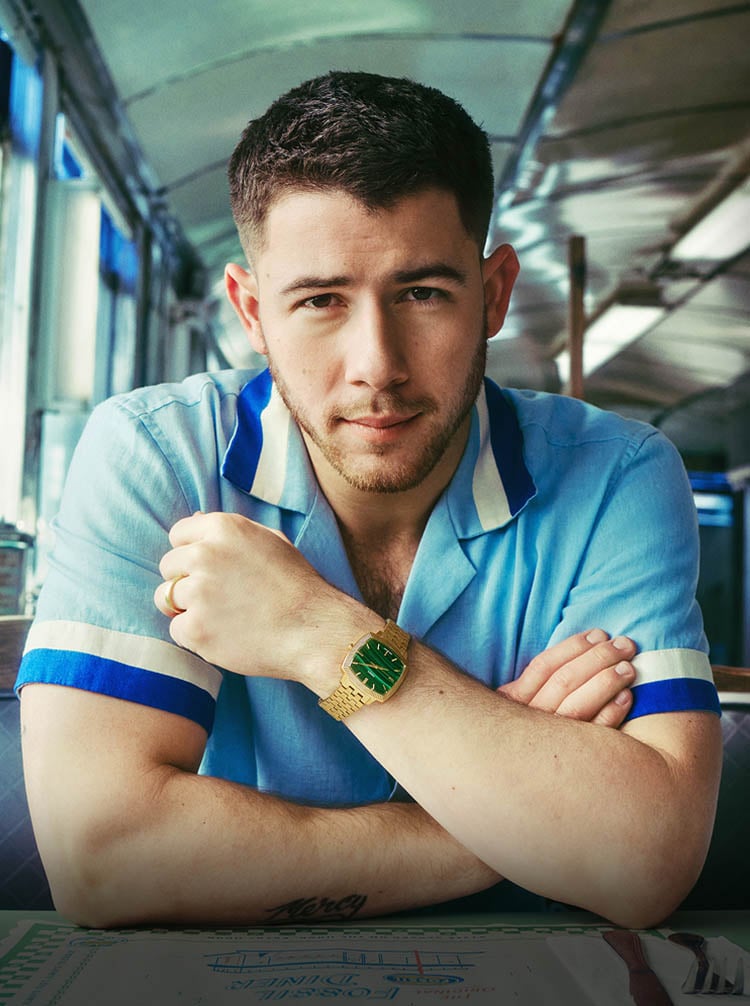 Nick Jonas est assis dans une banquette de diner, portant la montre Machine Luxe ton or avec un cadran vert en malachite.
