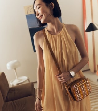 A woman with chin-length black hair is wearing a breezy halter-neck dress and light brown, patterned crossbody handbag.