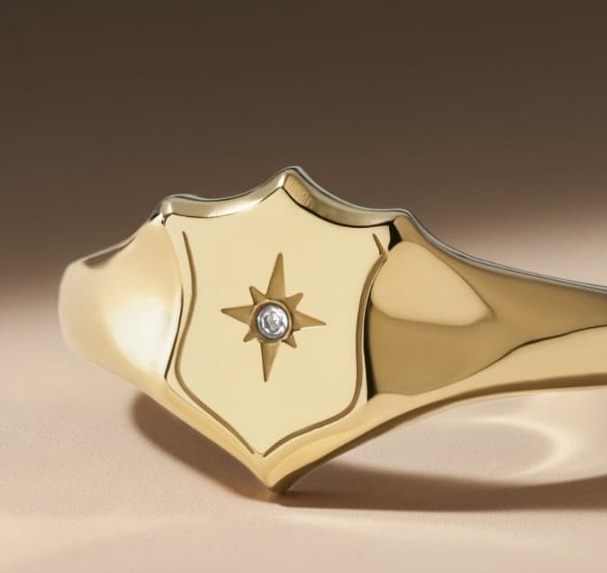 Una anillo de sello en tono dorado con la forma de nuestro escudo emblemático y un detalle de cristal.