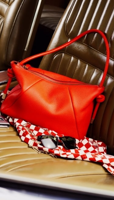 A bright red shoulder bag sitting on a chair with a red and white checkered scarf and sunglasses.