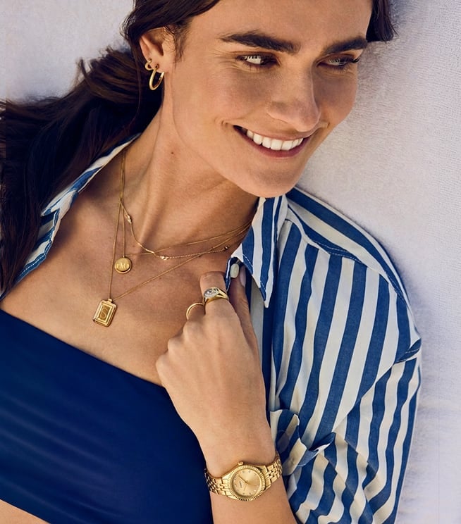 A woman lies on a lounge chair wearing a blue and white striped top, gold-toned Fossil Raquel jewellery, and a gold-toned Scarlette watch.