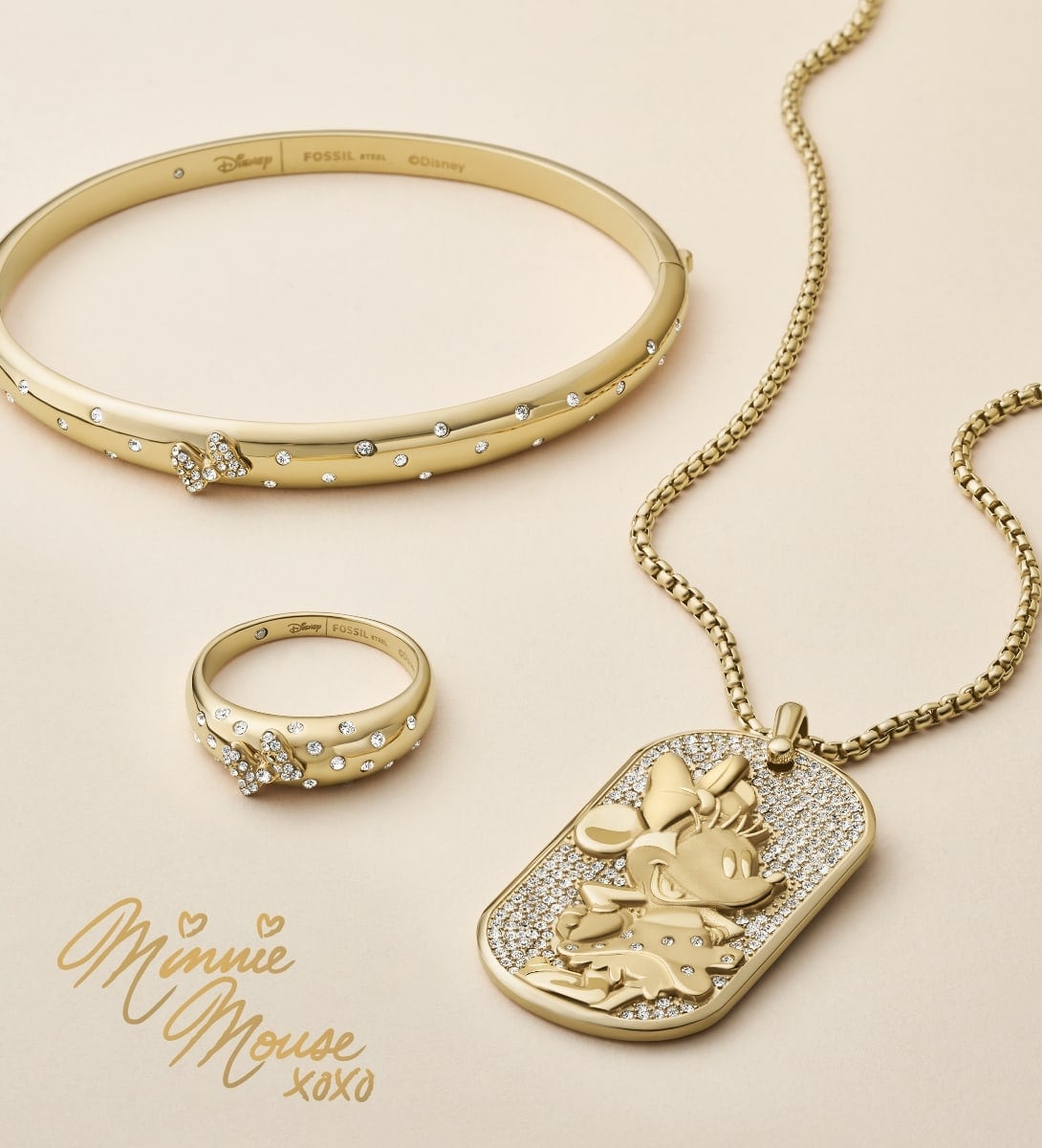 Three pieces of gold-tone Minnie Mouse jewelry, each accented with sparkling crystals to resemble Minnie's signature bow and polka dots. A bangle bracelet, ring and dog tag-style pendant necklace are artfully arranged on a cream-colored background. Minnie's signature is written in gold, along with hearts, x and o marks.