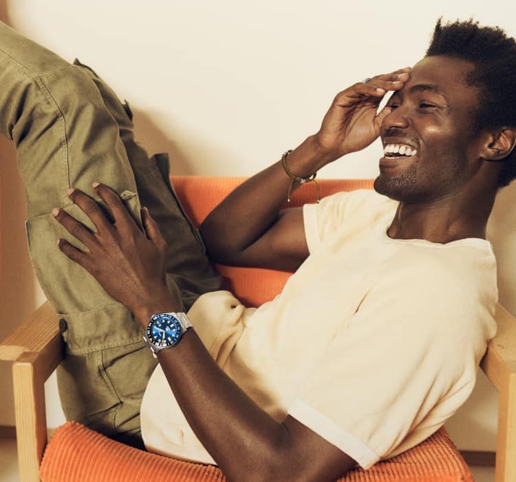 A man sits in a chair. He's wearing a Fossil Blue GMT watch: a stainless steel bracelet watch with a blue dial featuring geometric indices.