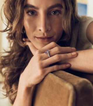 A woman with long wavy brown hair leans over a chair with her hands crossed, wearing a silver-toned ring.
