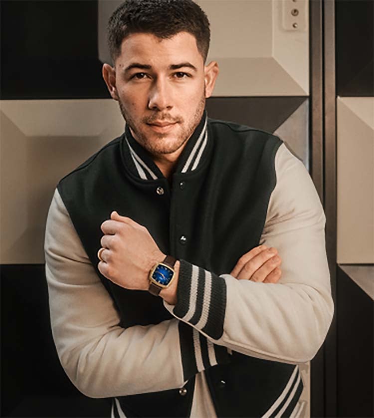 Nick Jonas leans against lockers at a bowling alley wearing a varsity jacket and the blue vignette Machine Luxe watch with a brown croc-embossed leather strap.