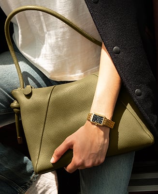 Eine Frauenhand, die eine Tasche aus ockerfarbenem Leder hält, mit einer Uhr Raquel mit goldfarbenem Finish und schwarzem Zifferblatt .