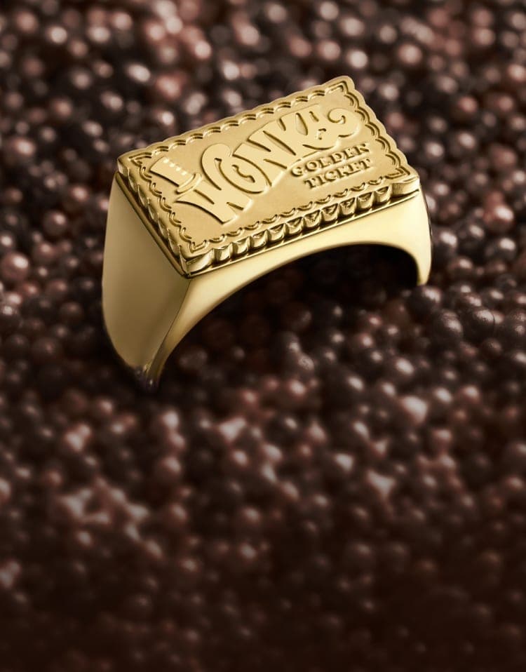 「金のチケット」のようなデザインのゴールドトーンのシグネットリングが、散りばめられたチョコレートに浸されている。