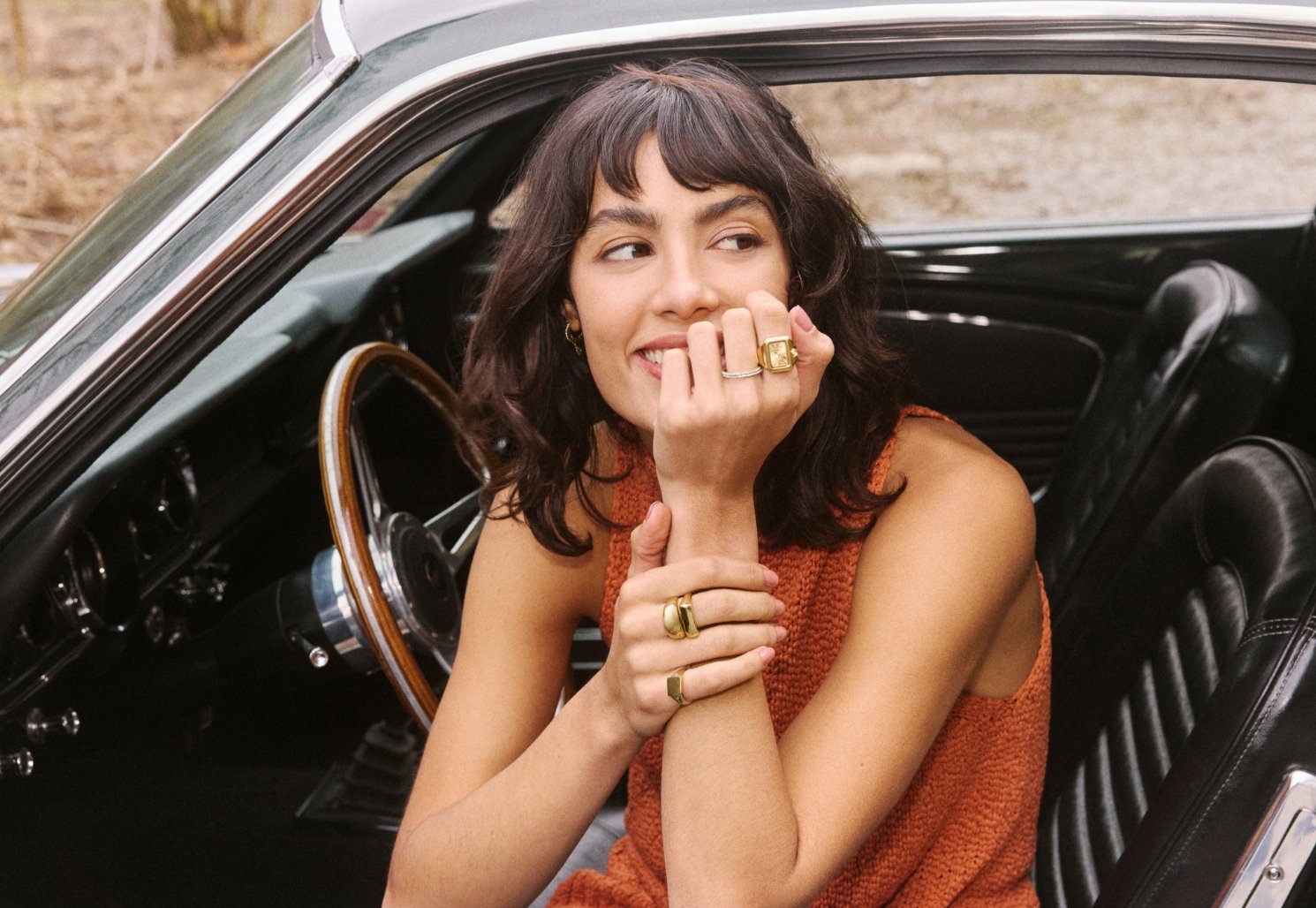 A woman sitting inside a car and wearing the Raquel watch ring and several gold-tone pieces of Fossil jewelry.