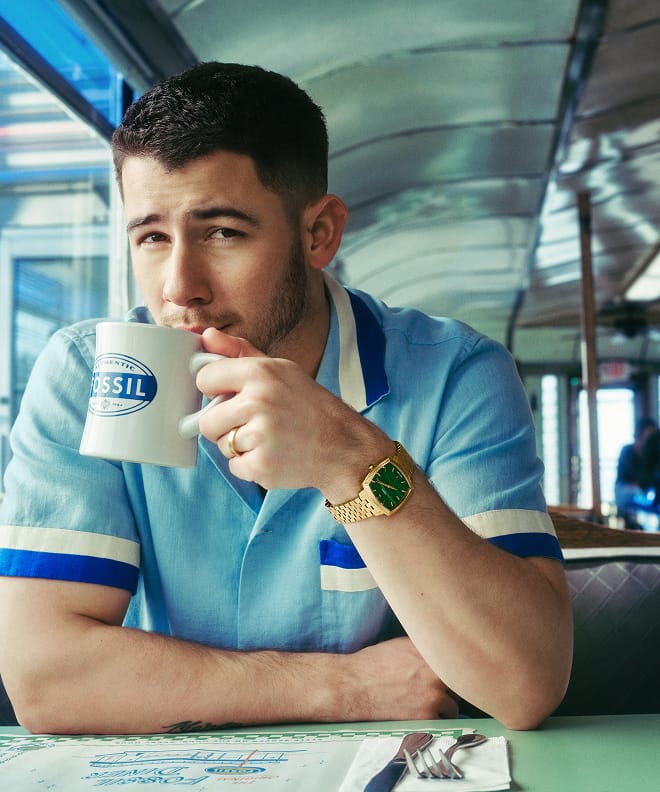 Nick Jonas sitting in a diner, drinking from a Fossil coffee mug and wearing the gold-toned Machine Luxe watch with a green malachite dial.