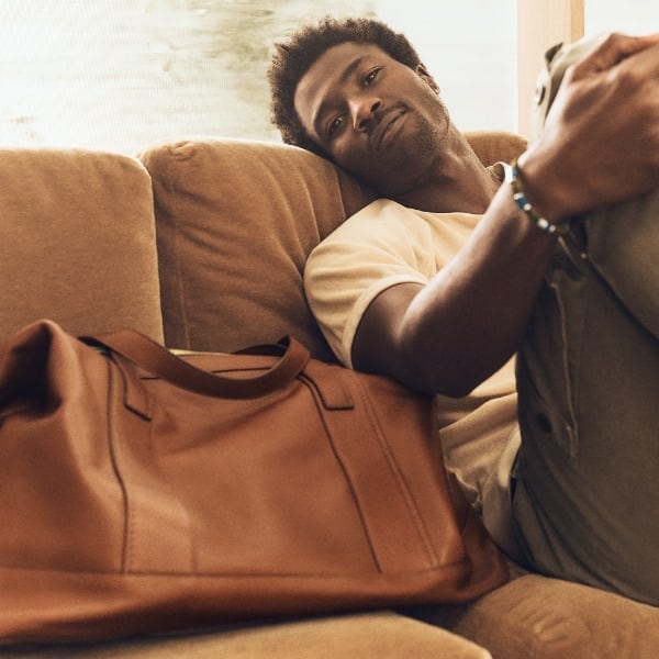 A man sitting on the sofa with the brown leather Raeford duffle next to him.