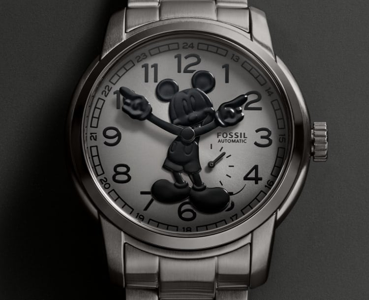 A silver-tone watch with a gradient dark grey dial featuring Disney’s Mickey Mouse. His silhouette is made to look like his shadow in glossy three-dimensional black. Mickey’s gloved hands rotate to tell the time. The watch is set against a coordinating off-white background.