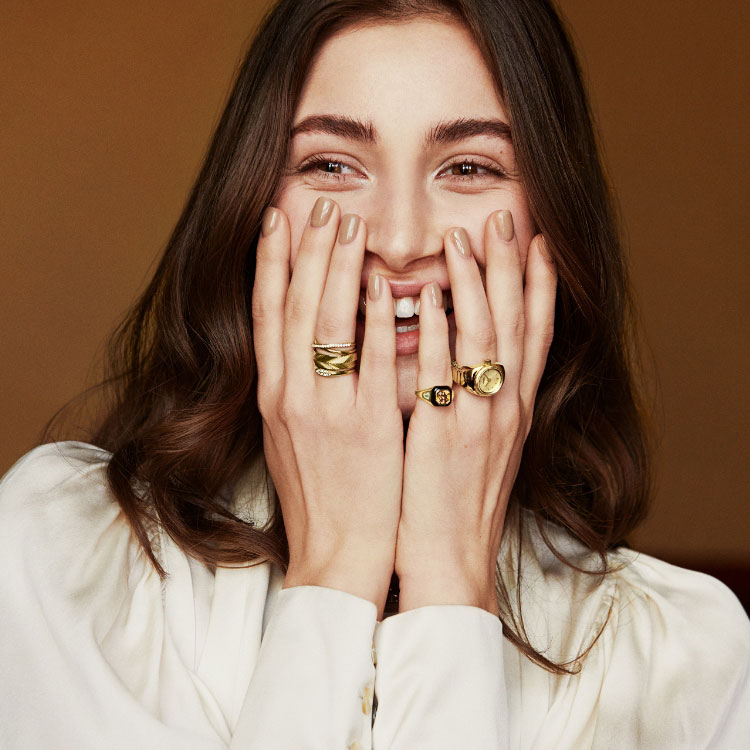 Imagen de una mujer que lleva puestos anillos dorados y se tapa la sonrisa. Imagen del anillo de reloj dorado con esfera de malaquita verde.