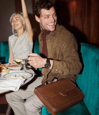 A man and a woman sitting together at a dinner table next to the brown leather Lennox messenger.