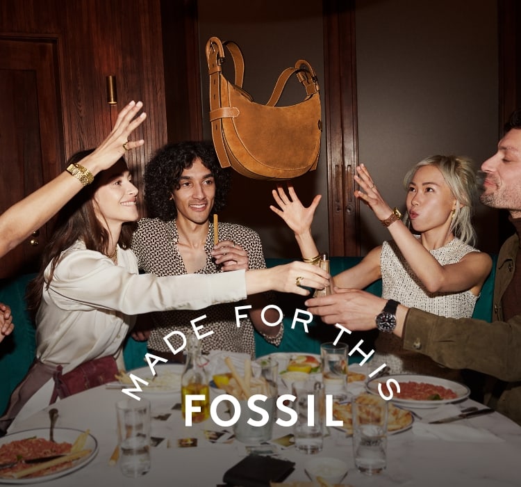 Un gruppo di amici seduti a tavola che lanciano in aria la borsa Harwell in pelle scamosciata marrone. Fossil, per chi ama la vita