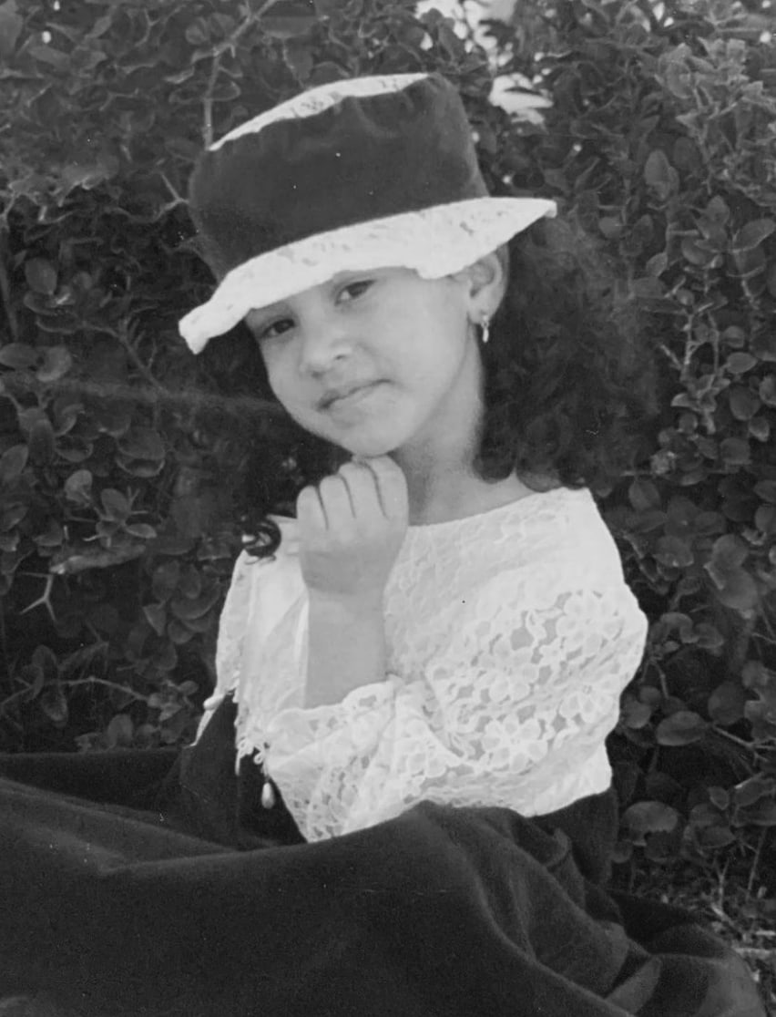 A gif image. The first photo shows Julissa Prado, smiling. Her curly hair is upswept and she's wearing the jewelry from this special-edition collection, complented by a light sage green strapless top. The second image shows a black-and-white photo of Julissa as a young girl, wearing a white lace brimmed hat accented with a dark ribbon and matching dress. 