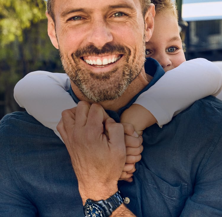 A smiling dad wearing the Fossil Townsman automatic watch. His son drapes his arms over his shoulder.
