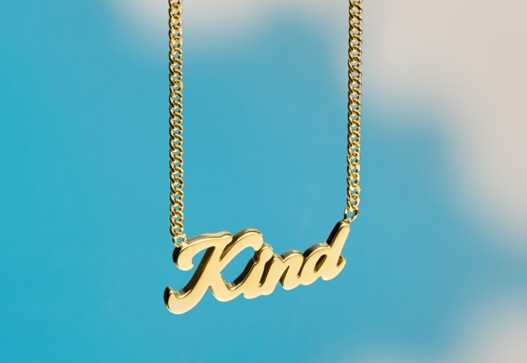 A blue sky background with clouds and a gold-tone La La Land x Fossil necklace that reads Kind.
