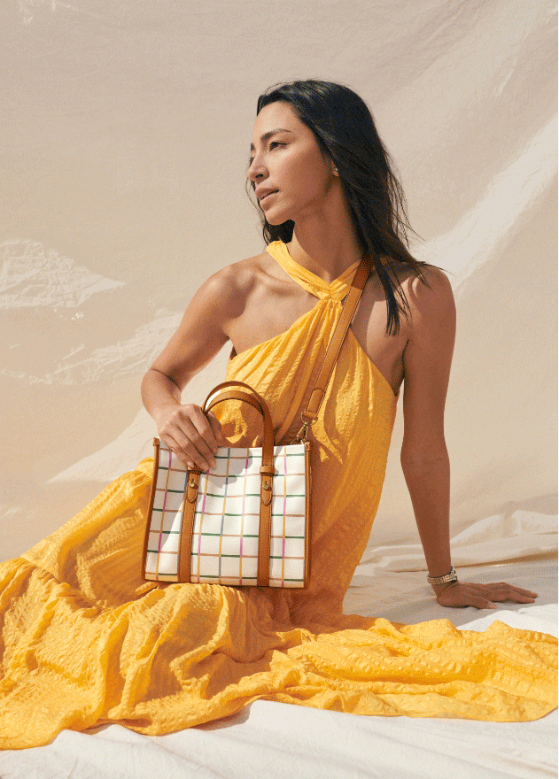  A woman sitting down, looking into the distance. She’s wearing a yellow sundress and holding a white handbag with tan handles and a rainbow grid pattern.