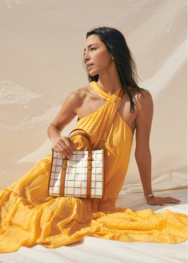 Ein sitzendes Model, das in die Ferne blickt. Es trägt ein gelbes Sommerkleid und hält eine weiße Tasche mit hellbraunen Henkeln und einem regenbogenfarbenen Gittermuster.