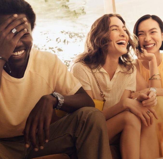 Drei Freunde (ein Mann und zwei Frauen), die zusammensitzen und lachen. Sie tragen luftige, beige Kleidung und modernen Schmuck mit gold- und silberfarbenen Finishs.