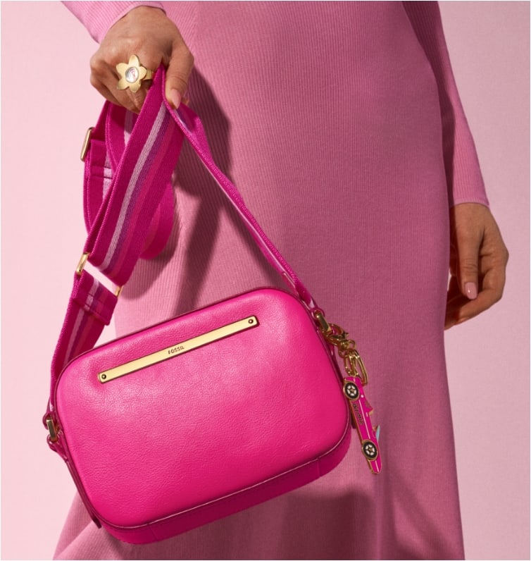 Une femme portant notre sac pour appareil photo Liza en édition limitée Barbie™ x Fossil en cuir pleine fleur rose emblématique avec une large bandoulière sportive rayée rose, style années 90.
