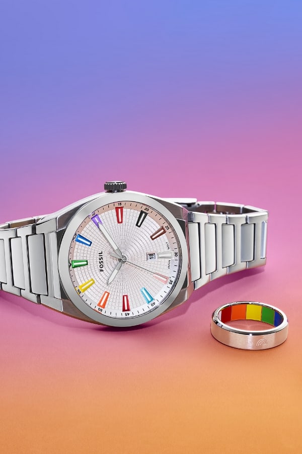 A silver watch and ring display rainbow accents that represent the colours of the Pride and Trans flags. They’re featured on a rainbow gradient background that transitions from purple to pink to orange.