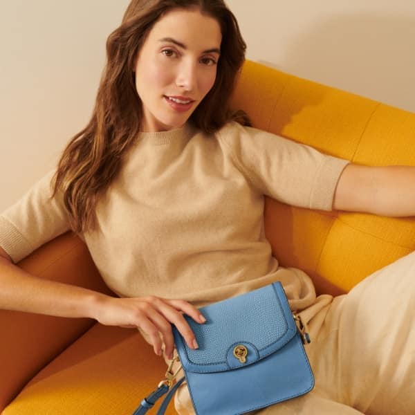A woman casually sitting on a yellow sofa. She’s wearing a beige short sleeve jumper and trousers, holding a small light blue crossbody handbag.
