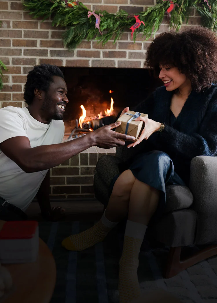 Two friends smiling, enjoying a cozy gift exchange in front of a fireplace. The image is framed in green and accented with holiday graphics, including drifts of snow and ornaments.