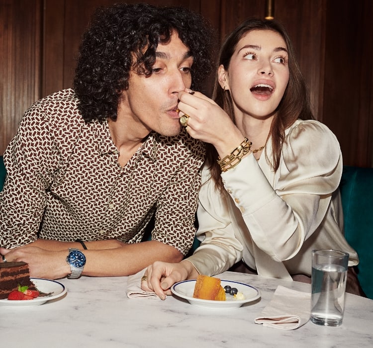 Un hombre y una mujer comiendo tarta. Ella lleva el anillo de reloj y joyas en tono dorado. Él lleva el reloj Fossil Blue GMT.