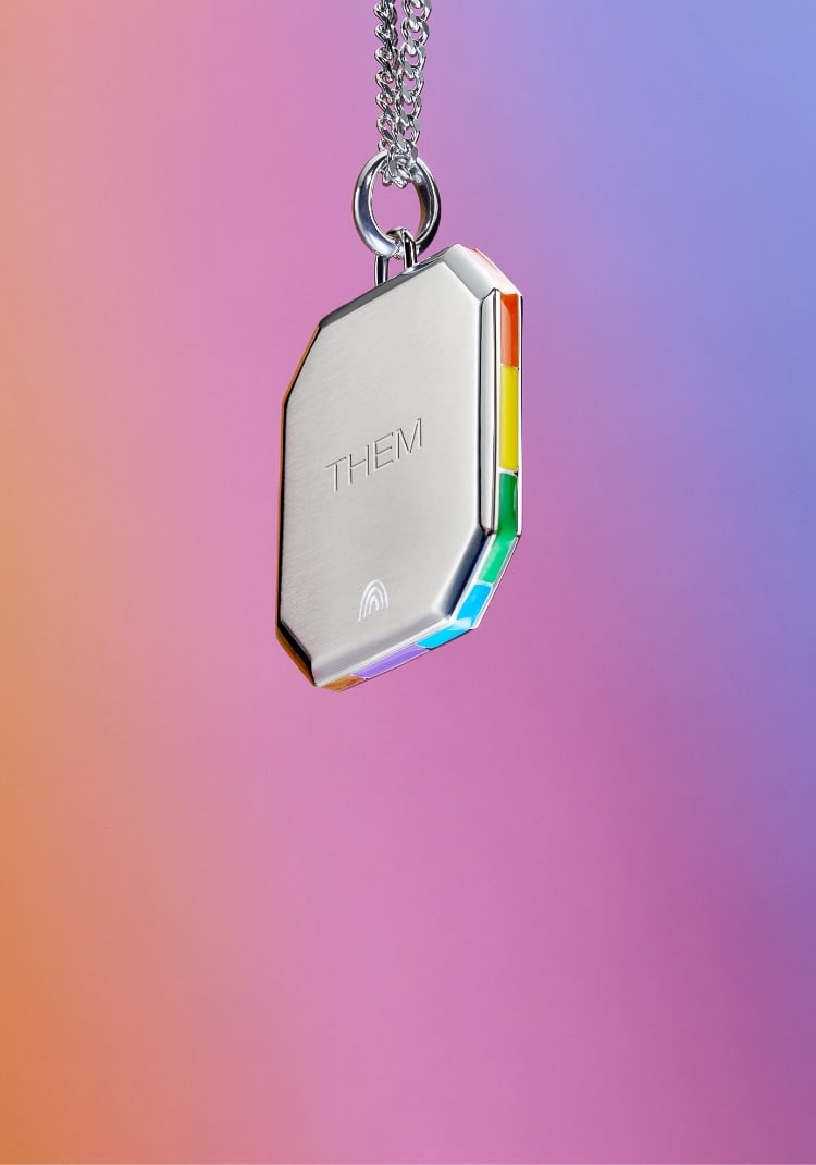 A silver geometric pendant necklace is suspended from a chain over a rainbow gradient background. The pendant is engraved with the pronoun Them and displays the colours of the Pride and Trans flags along the edges, as well as a small etched rainbow motif.