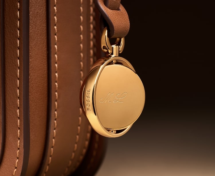 The gold-tone Legacy Charm hanging from the brown leather Lennox bag and engraved with ML. Overlaid with a sketch.